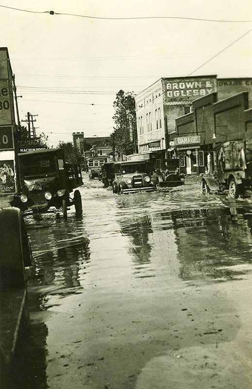 Cars driving on flooded town streets past storefront buildings
