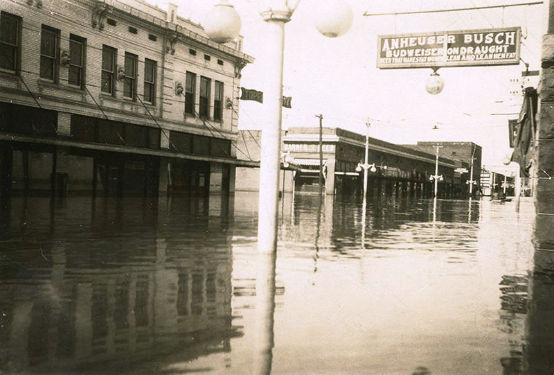 "Anheuser Busch" hanging sign over flooded town street with multistory buildings rising out of the water