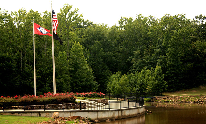 American and Arkansas flags on flagpoles near pond and trees