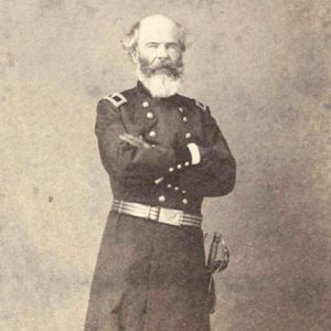 White man with long beard standing with arms crossed in military uniform with sword on his belt