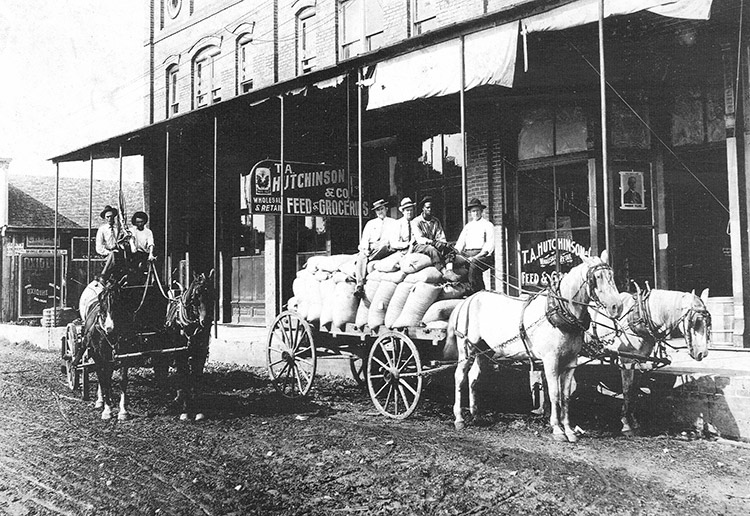 White men with horse drawn wagons on dirt road outside multistory feed and grocery store with covered porch