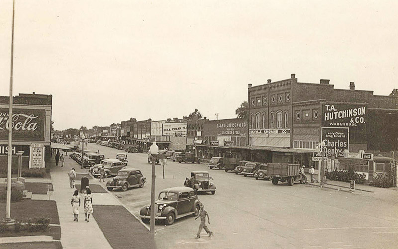 View of town street with parked cars and people walking along sidewalk in front of brick buildings