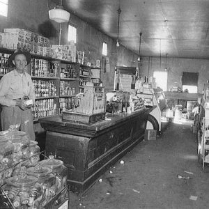 Older white man wearing a hat standing behind the counter inside general store
