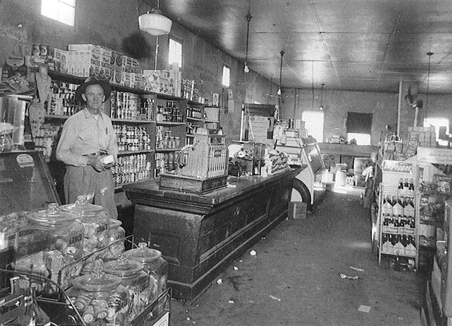 Older white man wearing a hat standing behind the counter inside general store