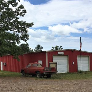 Red metal building with two garage bay doors flag pole and truck on dirt road