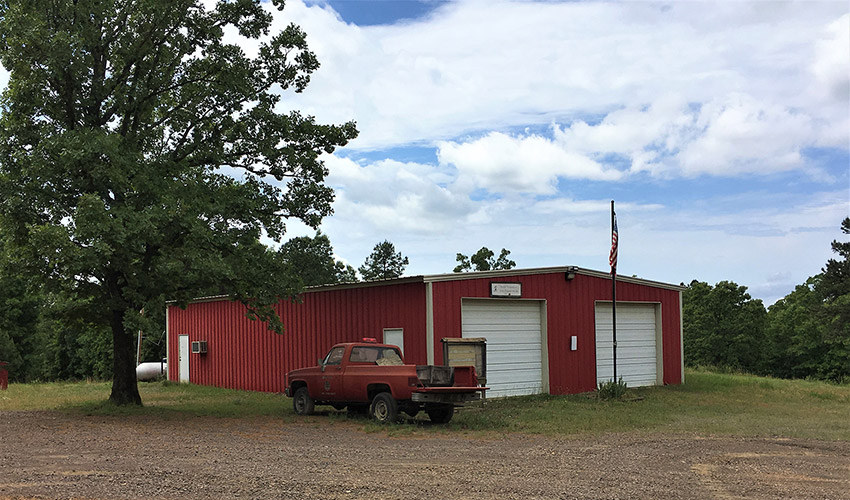 Red metal building with two garage bay doors flag pole and truck on dirt road