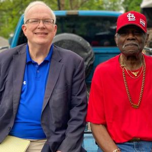 White man with glasses and African-American man in red hat and shirt with gold chain smiling in back of pickup truck