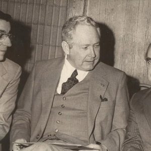 White man with glasses in suit and white man in suit both looking at third white man in suit with striped tie