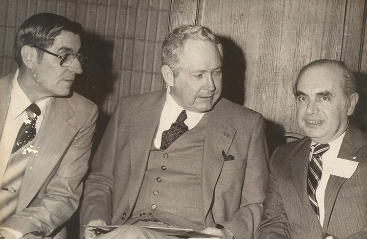 White man with glasses in suit and white man in suit both looking at third white man in suit with striped tie