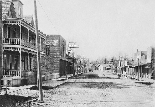 Multistory building with covered porch and balcony on dirt road with houses and storefronts in the background