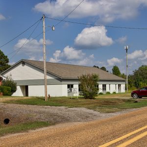 Single-story brick church building with steeple next to single-story building on two-lane road