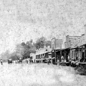People with horse-drawn wagons on town street with buildings on both sides