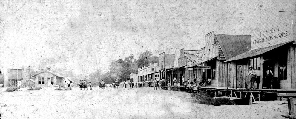 People with horse-drawn wagons on town street with buildings on both sides