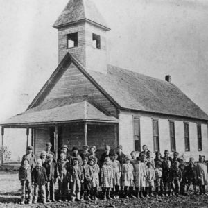 Group of white children and adults standing outside single-story building with covered porch and bell tower