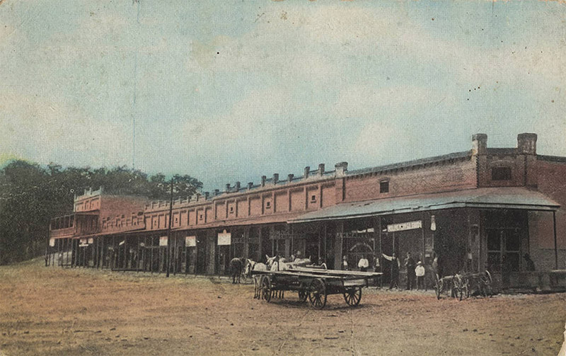 Multistory brick store fronts with covered walkway on dirt road with horse drawn wagon on it