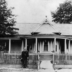 White man in suit standing outside house with round front porch and fence with gate