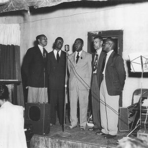 Five black men singing at microphone while black woman plays at piano