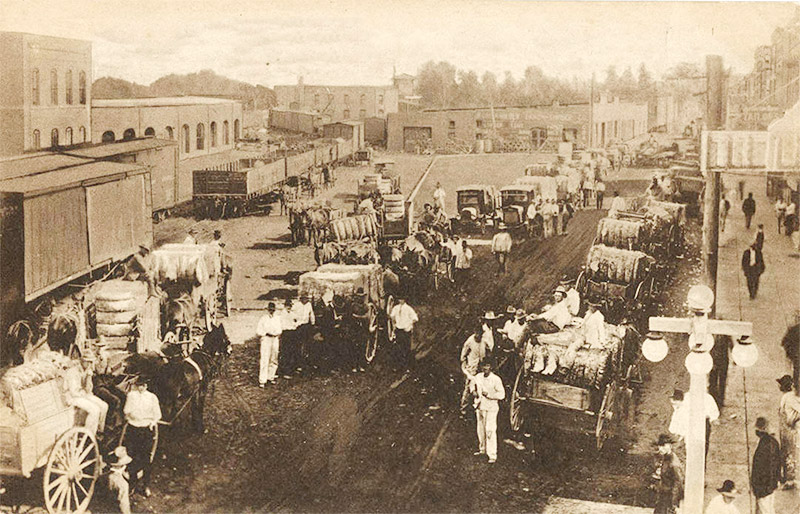 Horse drawn wagons loaded with cotton at rail yard with multistory buildings and storefronts in the background