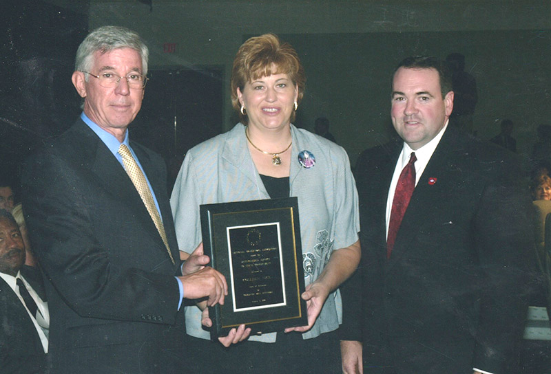 White woman holding plaque, standing between two white men in suits and ties