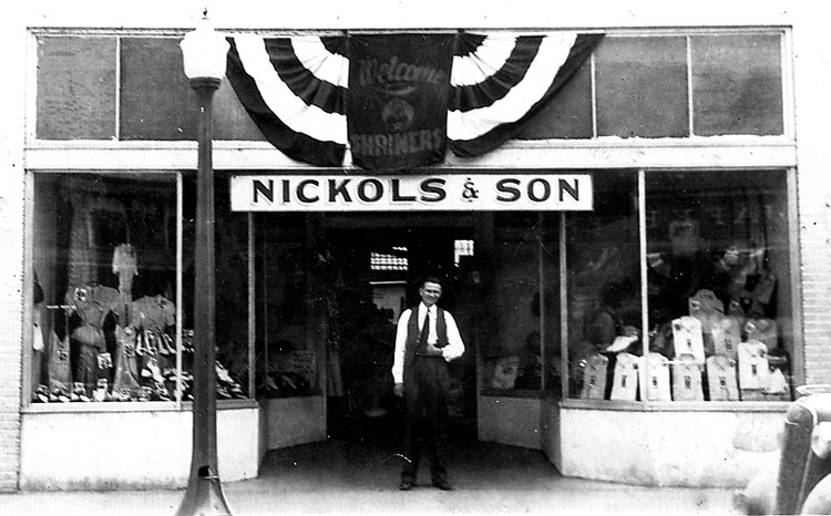 White man standing outside storefront with items in the windows and banners hanging above sign