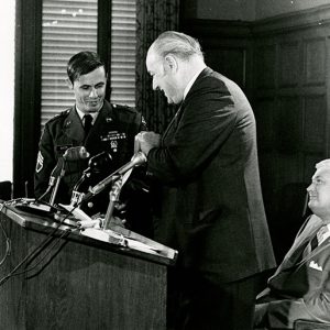 Older white man at lectern with microphones giving something to young white man in military uniform while another man sits behind them