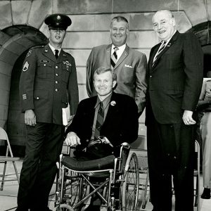 White man with no legs and a prosthetic arm in wheelchair with two white men in suits and one in a military uniform standing behind him