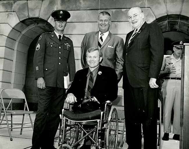 White man with no legs and a prosthetic arm in wheelchair with two white men in suits and one in a military uniform standing behind him