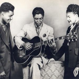 African-American man in suit and tie playing guitar with young African-American man and woman in suits watching him