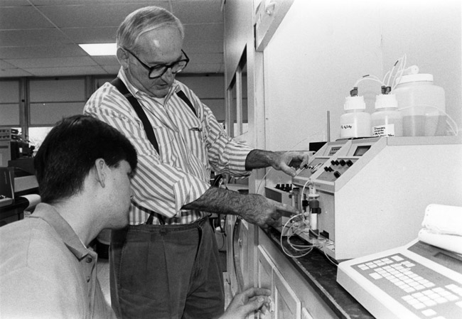 Old white man in suspenders teaching young white man to use lab equipment