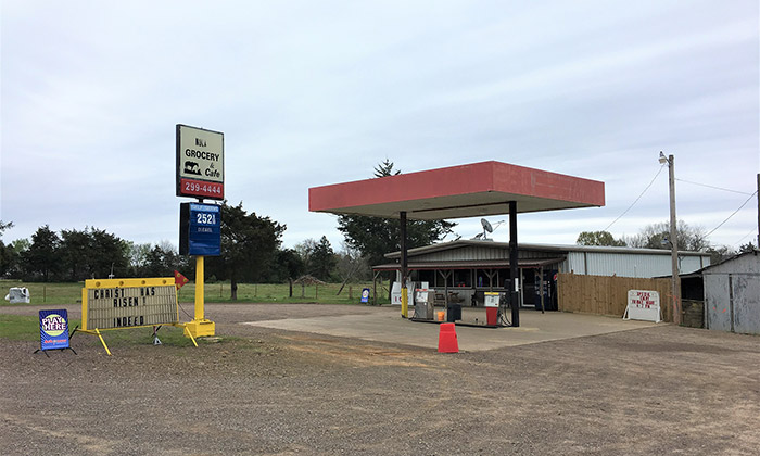 Single-story gas and grocery store with red canopy and signs on gravel parking lot