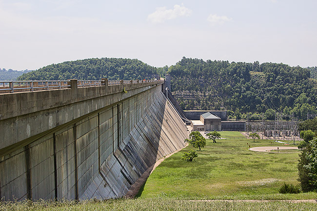 Concrete dam bridge with outbuildings and power station below it with tree-covered mountain in the background
