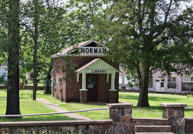 Narrow single-story "Norman Library" brick building with covered porch under trees in park with stone wall and steps in the foreground