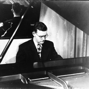 Young white man with glasses in suit and tie playing piano and reflected in mirror behind him