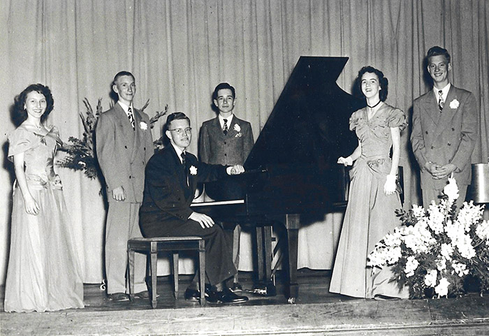 Young white men in suits and women in dresses on stage with young white man sitting at piano