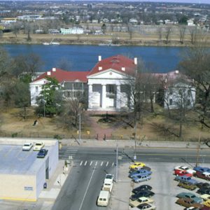 aerial view of Greek Revival structure with four front-facing columns and two wings with parking lot in foreground, river and city in background