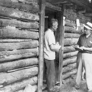 Two white men talking outside log cabin