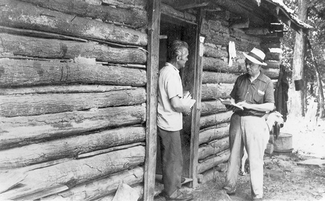 Two white men talking outside log cabin