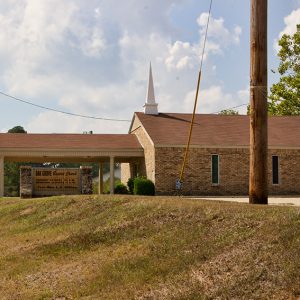 Side view of brick church building with steeple and covered entrance on parking lot with sign and telephone pole in the foreground
