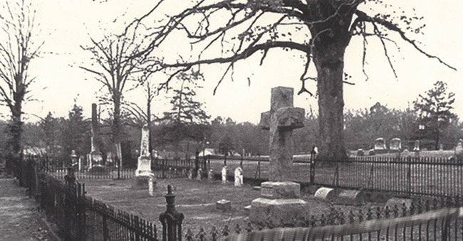 Tall monuments tombstones and cross grave marker surrounded by an iron fence and trees