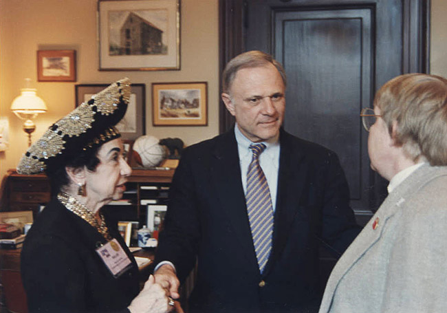 Older white woman speaking with two older white men in suits