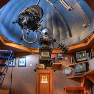 Interior of observatory with central telescope and ladder surrounded by walls featuring computers, monitors, clocks, and framed images