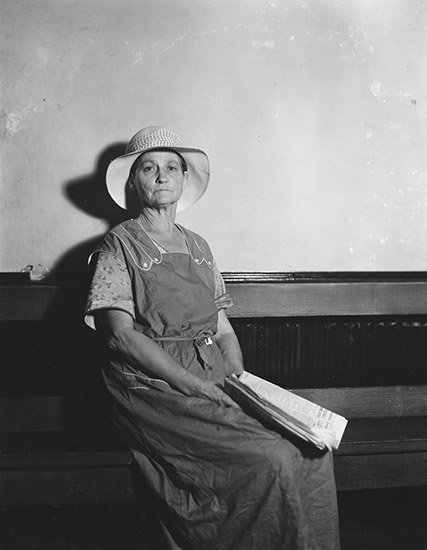 Old white woman sitting in hat and dress with newspaper on her lap