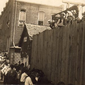 Brick building with men at gallows visible above wooden fence with crowd watching from the street