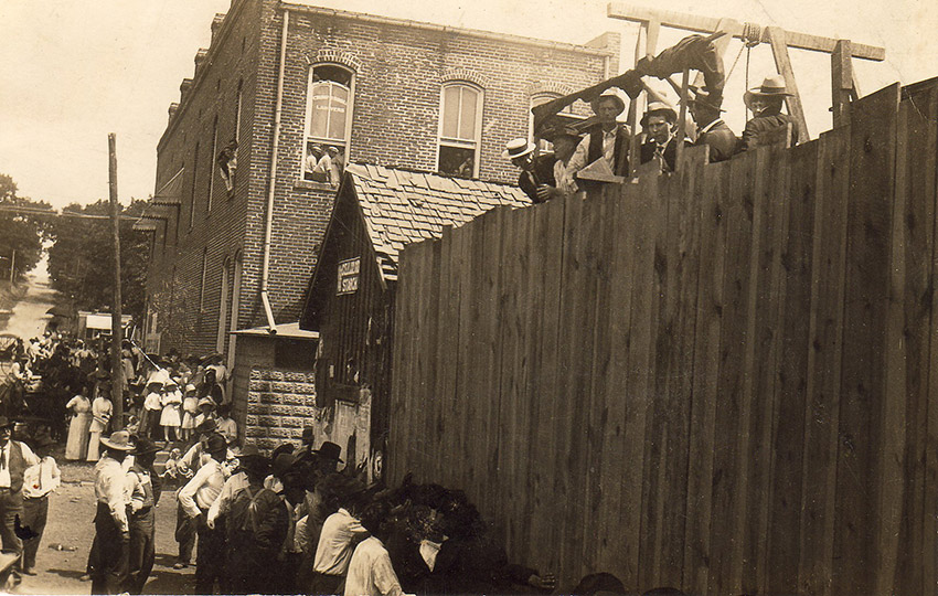 Brick building with men at gallows visible above wooden fence with crowd watching from the street
