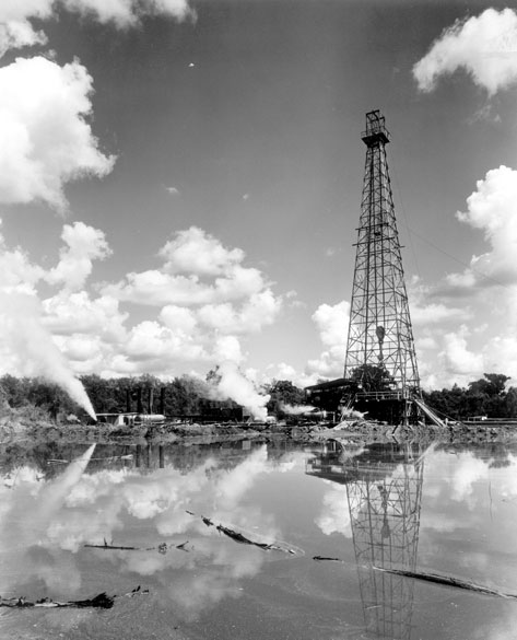 Oil derrick tower near body of water with sky reflected in the water