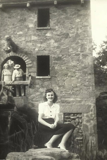 White woman sitting on rock wall with white couple in doorway of two-story mill building behind her