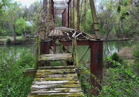 View looking across abandoned steel truss bridge over river with broken overgrown wooden landing in the foreground