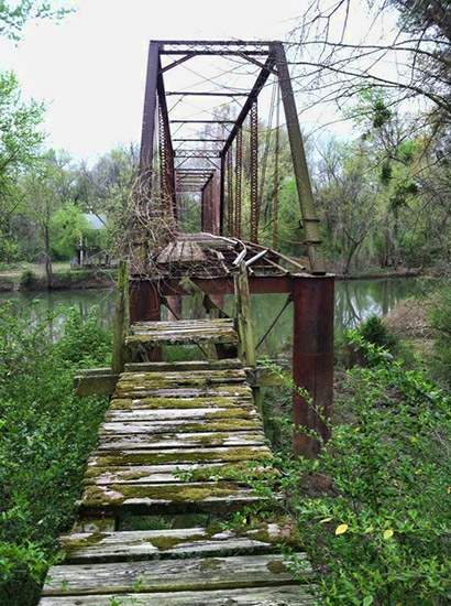View looking across abandoned steel truss bridge over river with broken overgrown wooden landing in the foreground