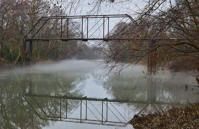 Steel truss bridge reflected in foggy river with trees on both sides
