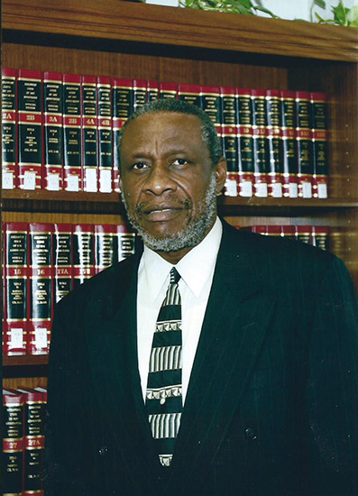 African-American man with graying beard in suit and tie standing at bookshelf
